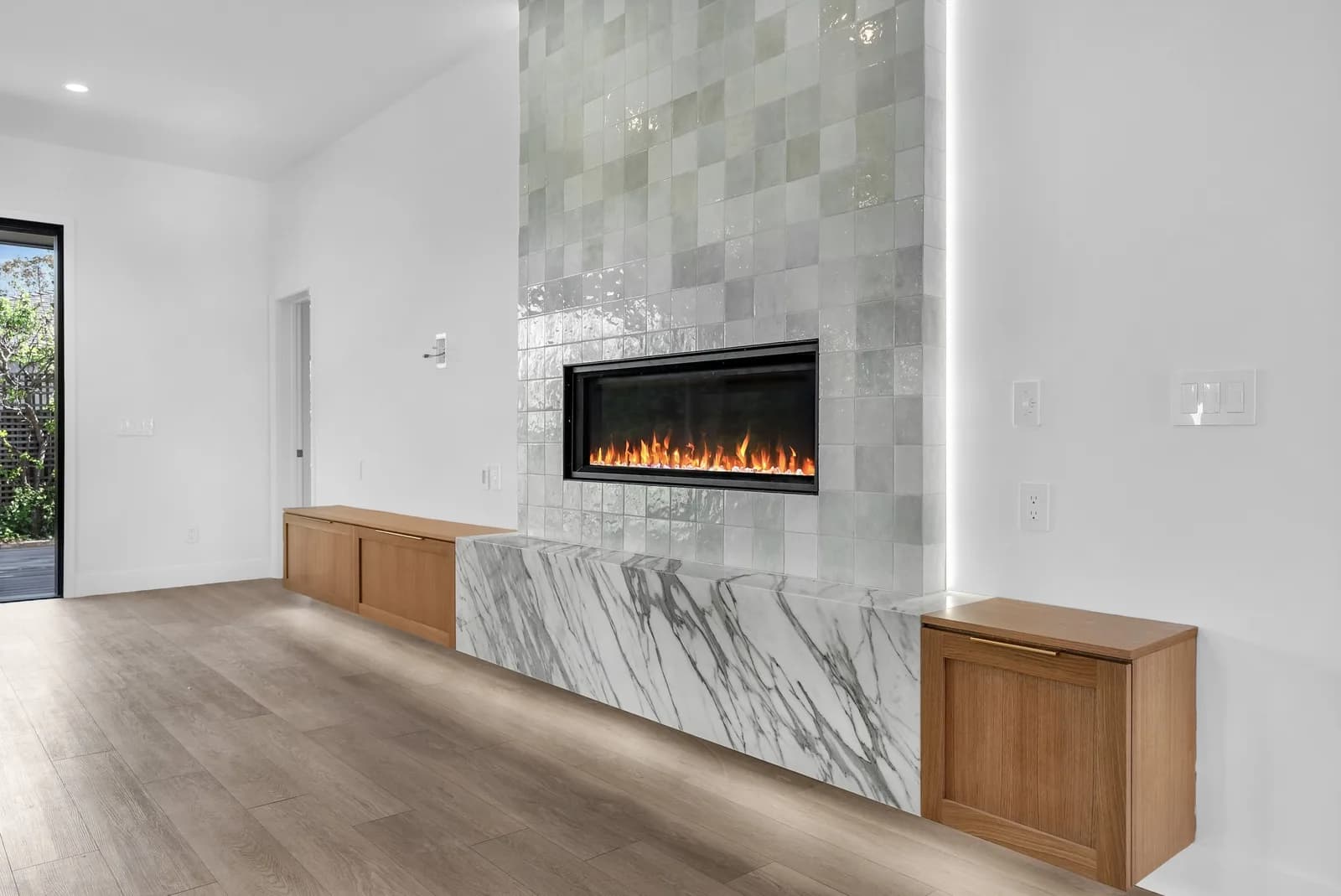 Fireplace wall in a finished living room by a network contractor: glazed sage-gray tile around a linear fireplace, a marble hearth bench and floating oak cabinets.