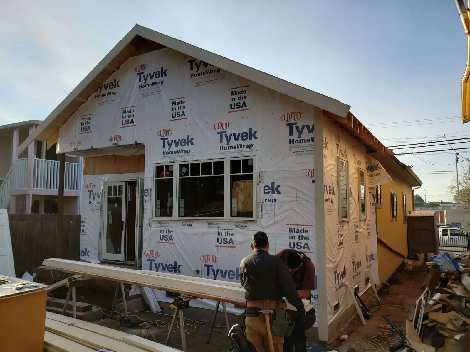 An active Bay Area job site during a home addition, framed and wrapped in housewrap, with two tradesmen working at a sawhorse out front.