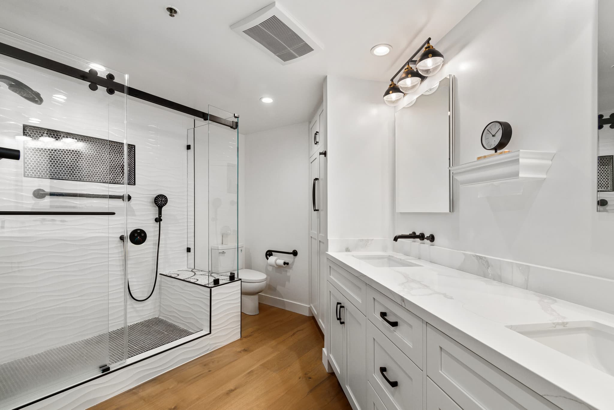 Remodeled primary bathroom with sculpted white wave tile, black-framed glass shower, and double quartz vanity